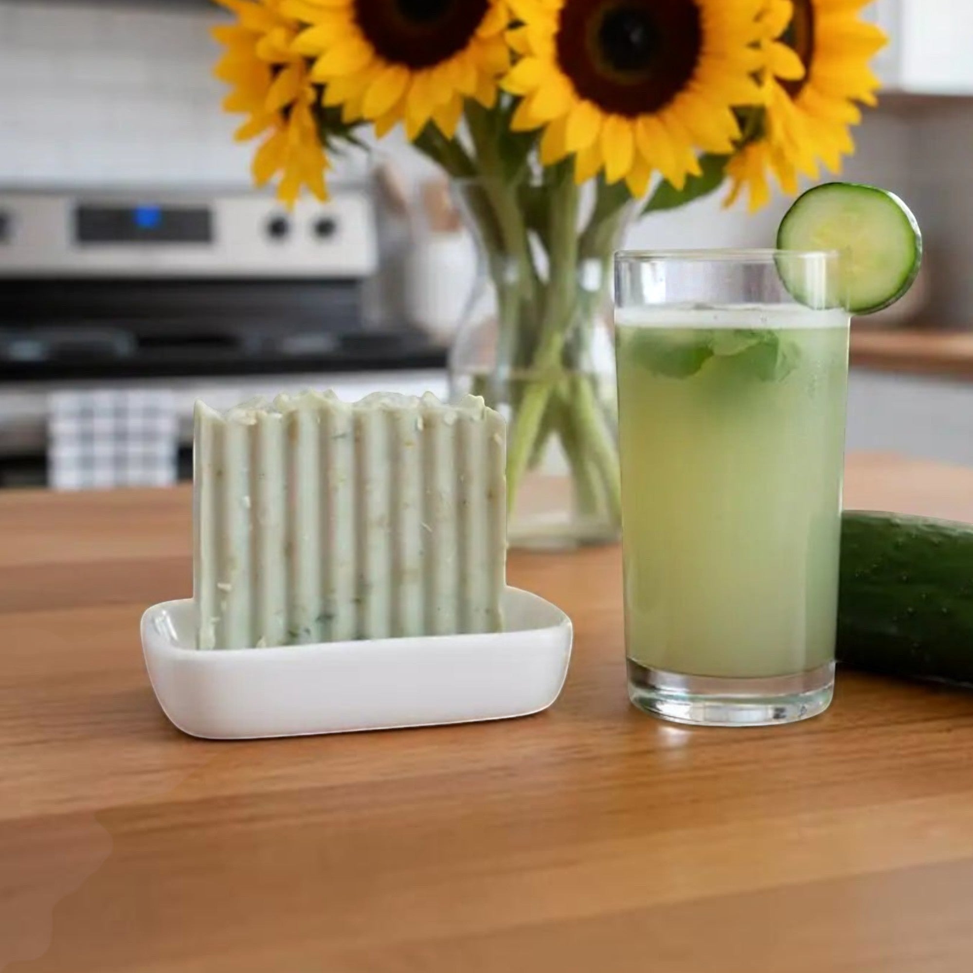Cucumber Bliss handmade soap on a white dish beside cucumber water and sunflowers in a bright spa inspired setting.